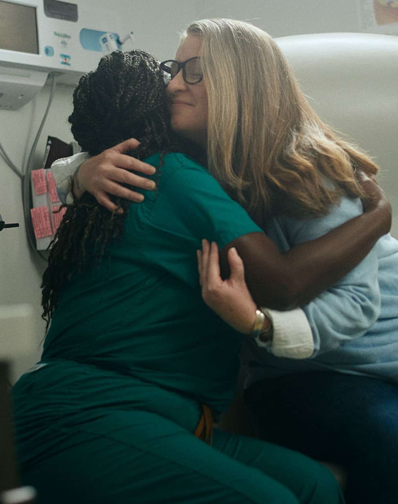 Two women embracing indoors, one in scrubs and the other in casual clothing, in what appears to be a healthcare setting.