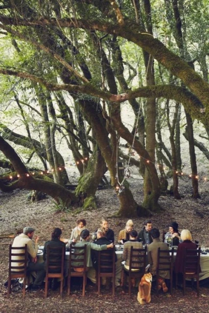 A group of people gathered around a long table for an outdoor meal in a forest setting, featuring lush trees and a warm, inviting atmosphere.