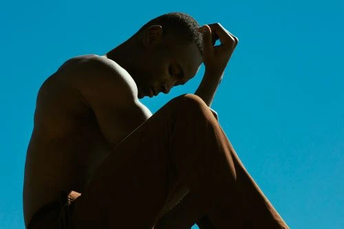 Dark-skinned adult male sitting with head down in a contemplative pose against a vivid blue background in an eye-level medium shot.