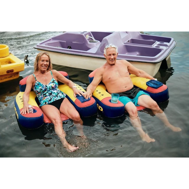 Couple relaxes on inflatable lounges in the water with a pedal boat in the background, enjoying a sunny day on the lake.