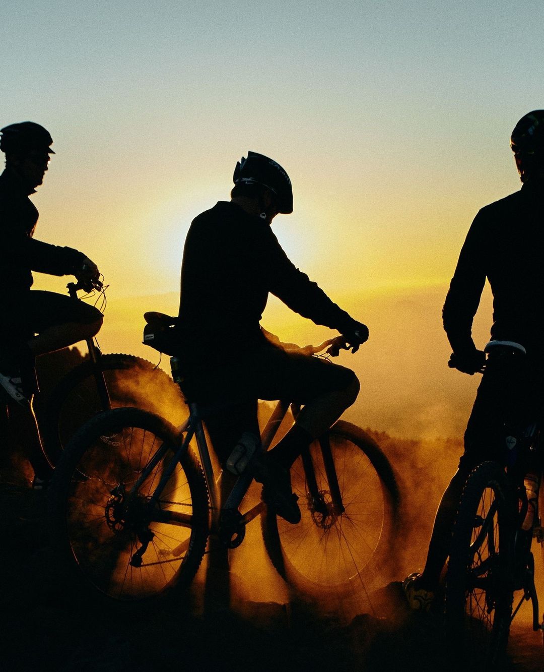 Three silhouetted cyclists biking at sunset, wearing helmets, riding on a dirt trail, with golden sky, creating a sense of adventure and freedom.