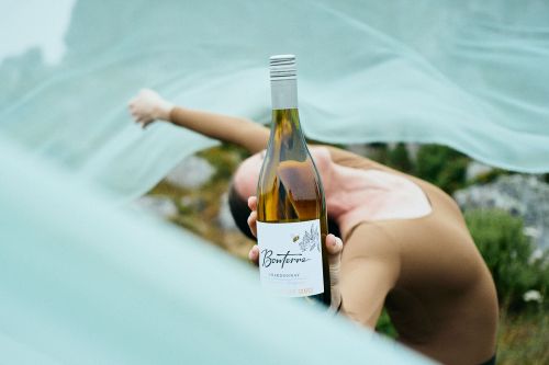 Wine bottle close-up held by person against blurred outdoor background with flowing fabric, promoting Bonterra Chardonnay.