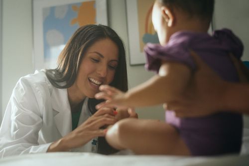 A female doctor smiles while interacting with a baby patient sitting on a medical examination table in a brightly lit clinic setting.