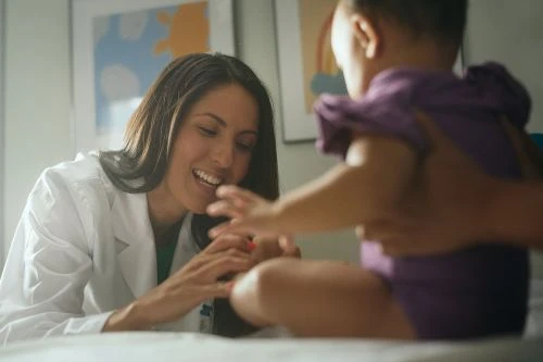 A female doctor smiles while interacting with a baby patient sitting on a medical examination table in a brightly lit clinic setting.