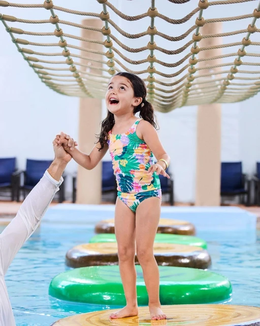 A joyful young girl in a colorful swimsuit stands on a floating platform in a pool, holding hands with an adult in an indoor waterpark.