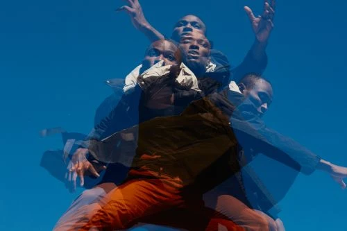 African-American man in orange pants and a black jacket, multiple exposures against a bright blue sky.