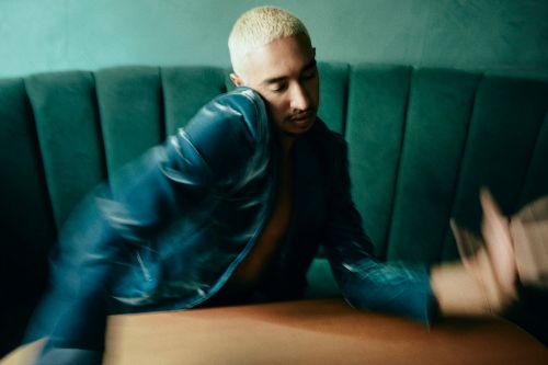 Stylish man with platinum blonde hair, wearing a navy blue jacket, sits against a dark green tufted booth with a blurred foreground.
