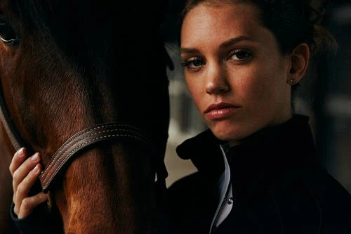 Close-up studio shot of a young woman with dark hair wearing a black jacket next to a brown horse with a leather bridle.