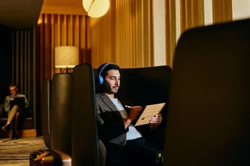 Adult man in a booth wearing headphones, using a tablet with a stylus in a modern lounge area with warm lighting and vertical striped walls.