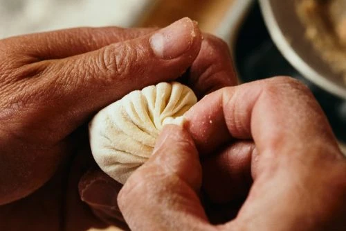 Close-up of human hands delicately shaping a dumpling, showcasing intricate folds and attention to detail in food preparation.