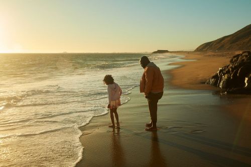 Girl and adult stand at water's edge on sandy beach at sunset with ocean and distant land, creating a scenic, reflective moment.