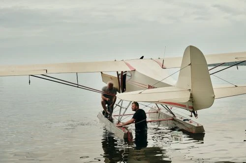 Two adult men stand on a seaplane's floats in the water, preparing for takeoff on a cloudy day, with the plane's wing and tail visible.