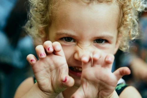 Close-up of a young child with curly hair making a playful 'claw' gesture with their hands toward the camera, creating a cute and mischievous moment.