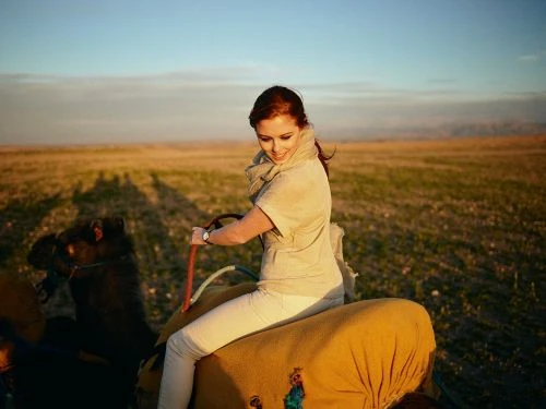 Woman with red hair rides a camel across a grassy field beneath a blue sky, wearing neutral clothing and white pants.