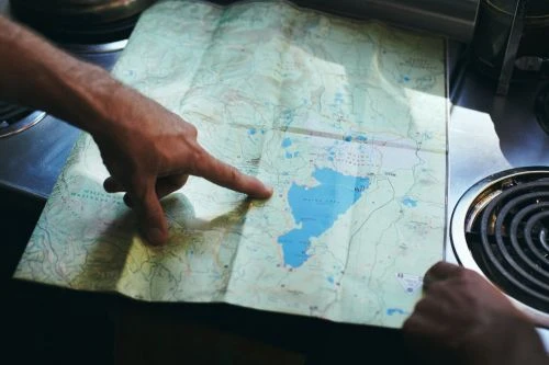 Close-up of a person's hands pointing to locations on a topographic map with mountains and blue lakes. Cooking stove in the background.