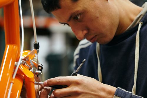 Close-up of a man in a hooded sweatshirt working on an orange bicycle with tools in an indoor setting, with blurred background elements.