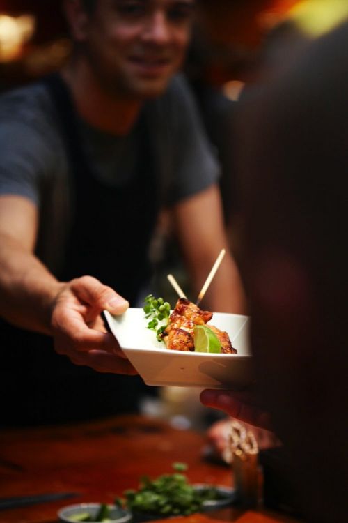 Man in black apron serves plate of chicken skewers with lime garnish to an unseen person indoors under warm lighting.