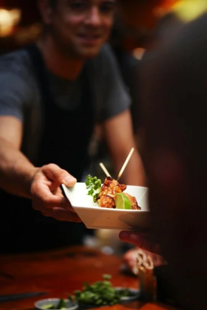 Man in black apron serves plate of chicken skewers with lime garnish to an unseen person indoors under warm lighting.