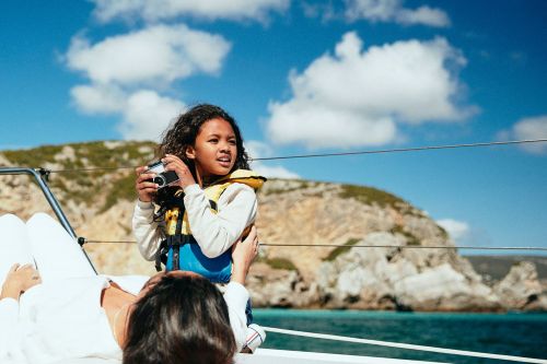 Child with curly hair and wearing a life jacket holds a camera on a boat with an adult in the foreground and an island in the background.