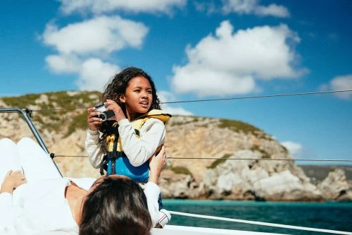 Child with curly hair and wearing a life jacket holds a camera on a boat with an adult in the foreground and an island in the background.