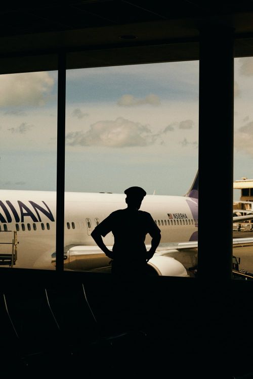 Silhouette of an adult person wearing a hat inside an airport terminal looking out at a Hawaiian Airlines airplane on a sunny day.