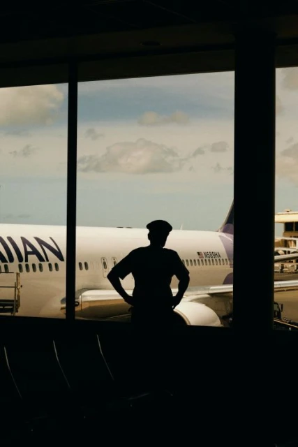 Silhouette of an adult person wearing a hat inside an airport terminal looking out at a Hawaiian Airlines airplane on a sunny day.