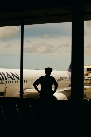 Silhouette of an adult person wearing a hat inside an airport terminal looking out at a Hawaiian Airlines airplane on a sunny day.