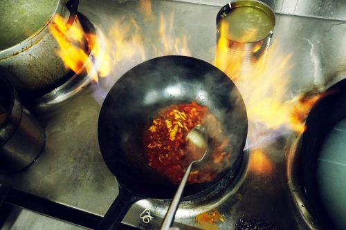 Overhead shot of a wok on a stove with food being cooked amid flames, with other cooking vessels visible in the background.