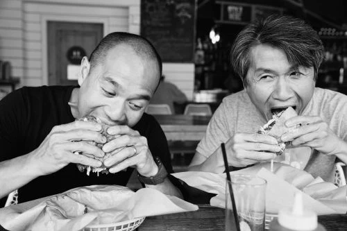 Two men are eating hamburgers at an indoor restaurant. The black and white photo shows them enjoying their meal with visible drinks and condiments.
