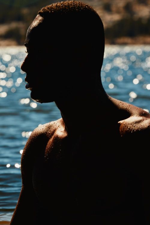 Silhouette of a muscular Black man emerging from blue water, back-lit by natural light, highlighting skin texture and the ripple effect on the water.