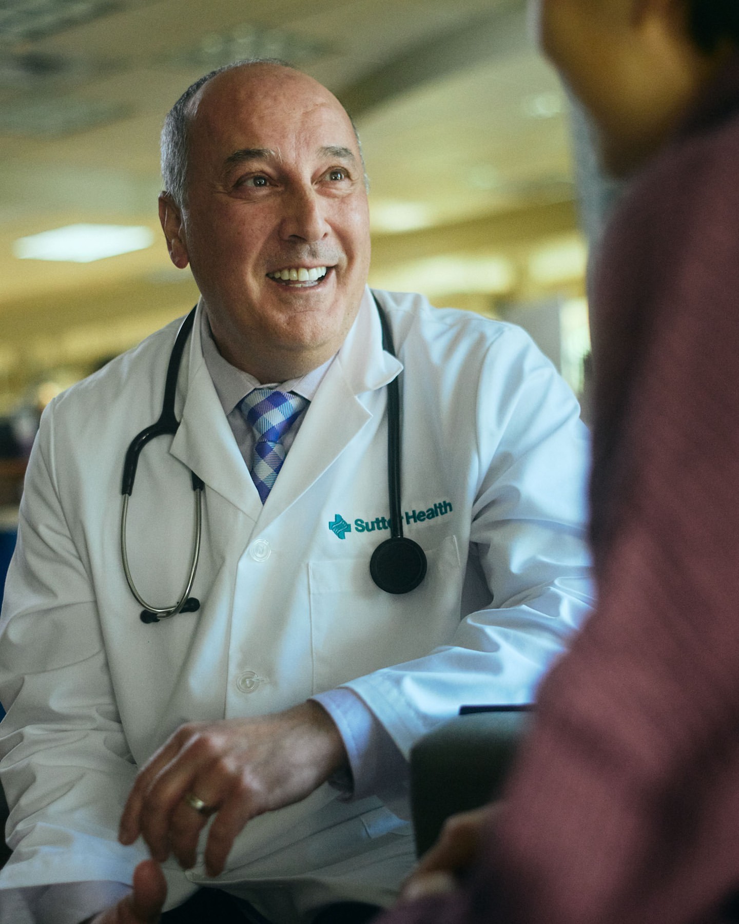 Friendly mature doctor with stethoscope and lab coat smiles at patient during a consultation in a bright, modern medical office.