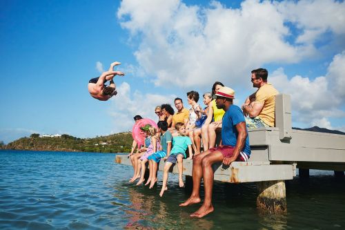 Group of people watching a man do a backflip into the water near a dock on a sunny day with blue sky and scattered clouds.