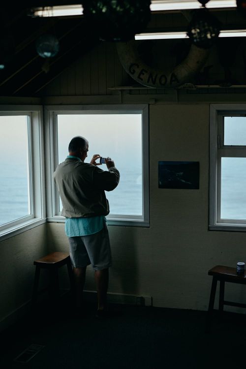 Adult male in shorts stands in a corner room with large windows, taking a picture of the water outside with his cell phone.