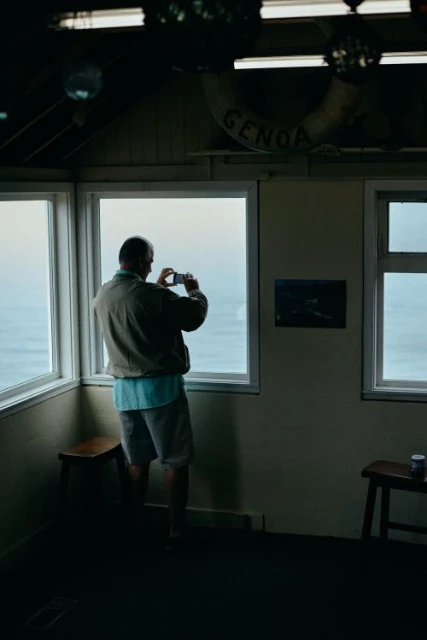 Adult male in shorts stands in a corner room with large windows, taking a picture of the water outside with his cell phone.