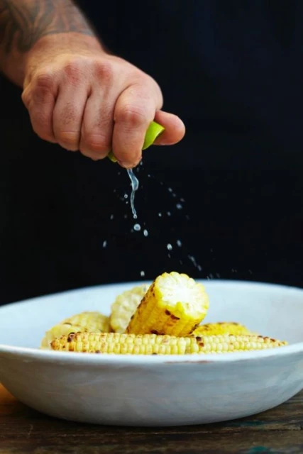 Tattooed hand squeezing lime juice over grilled corn on the cob in a white bowl, set against a dark backdrop, ready for a tasty meal.