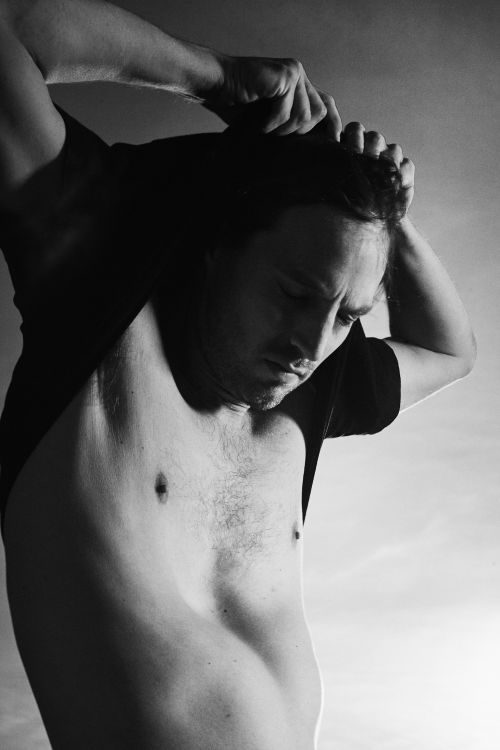 Monochromatic close-up of an adult man partially undressed, removing a dark t-shirt, showcasing torso against a bright, gradient background.