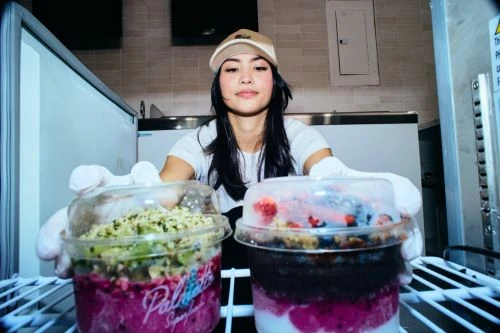 Asian woman in white gloves holds two smoothie bowls with fresh fruit in a bright indoor food service environment. Close-up, eye-level view.