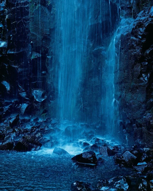 Dramatic view of a waterfall cascading over dark rocks into a pool of water, illuminated by blue-toned lighting.