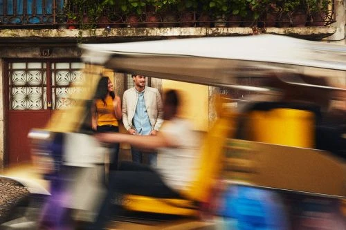 Couple in yellow and blue on urban street, historic building backdrop with lush green plants. Blurred motion street scene.