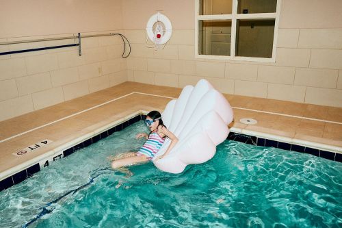 Young girl in a striped swimsuit and goggles floats on a large inflatable seashell in a blue-tiled indoor swimming pool.