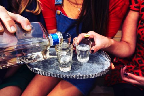 Person pouring clear liquid from a glass bottle into shot glasses on a silver tray, surrounded by three people wearing casual attire.