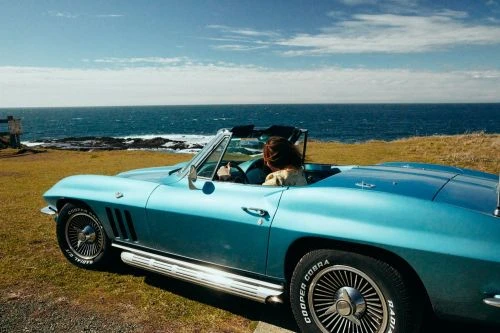 Woman driving a light blue vintage convertible car along a coastal road with the ocean and sky visible in the background.