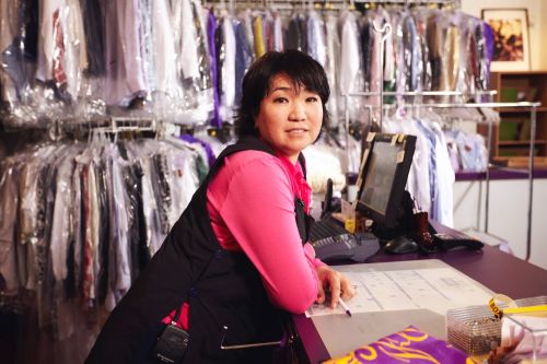 Friendly dry cleaning service worker standing behind the counter in front of rows of cleaned and bagged clothes ready for pickup.