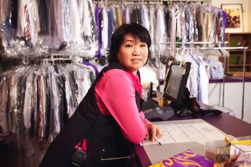 Friendly dry cleaning service worker standing behind the counter in front of rows of cleaned and bagged clothes ready for pickup.