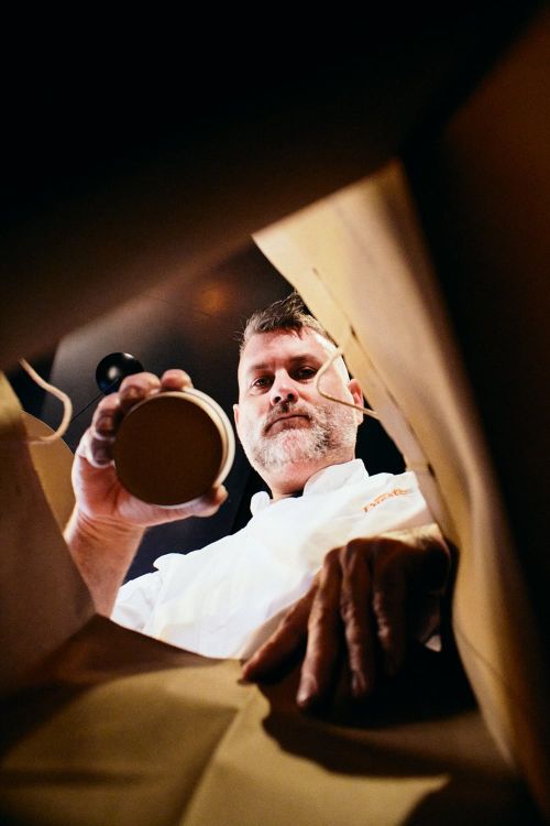 Eye-level view into a brown paper bag featuring an adult male chef with facial hair in a white chef's coat holding round container.