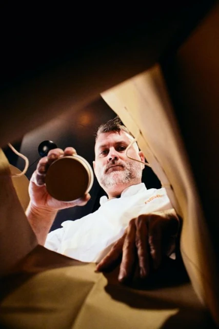 Eye-level view into a brown paper bag featuring an adult male chef with facial hair in a white chef's coat holding round container.