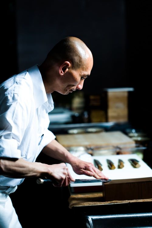 Asian chef wearing white uniform, preparing food on a counter with kitchen knife and ingredients in a dimly lit kitchen setting.