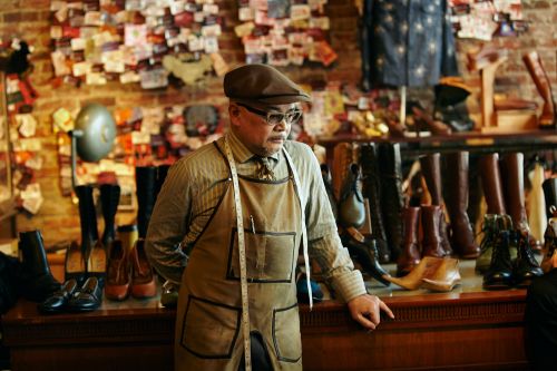 Craftsman in cap and apron stands in his shoe store, surrounded by a collection of boots and shoes displayed on wooden shelving.