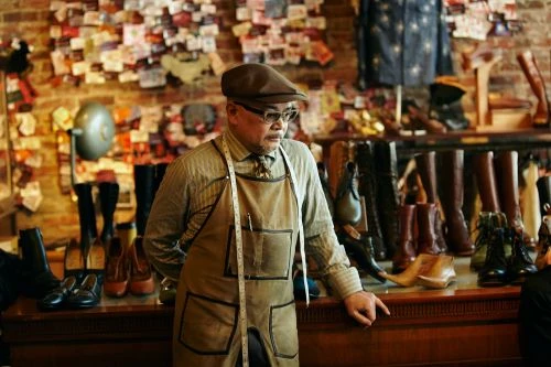 Craftsman in cap and apron stands in his shoe store, surrounded by a collection of boots and shoes displayed on wooden shelving.