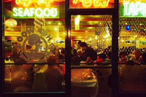 Busy Asian restaurant interior at night, seen through front window, with people dining, neon signs, and bright lights.